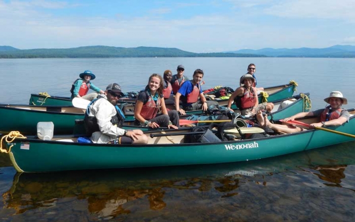 A group of canoes float near each other on calm water. The young people in the boats smile at the camera. In the distance there is a mountainous landscape.